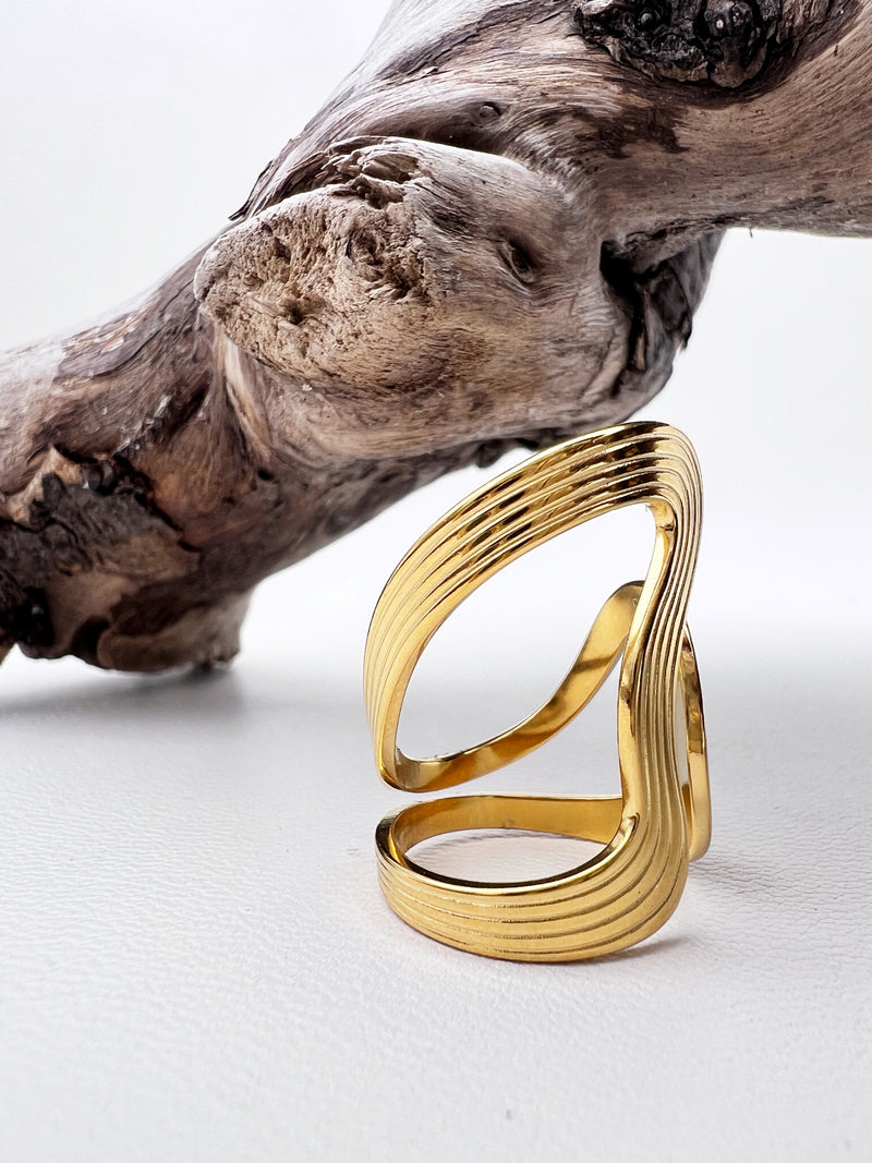 Gold wave ring on a white surface with a piece of driftwood.