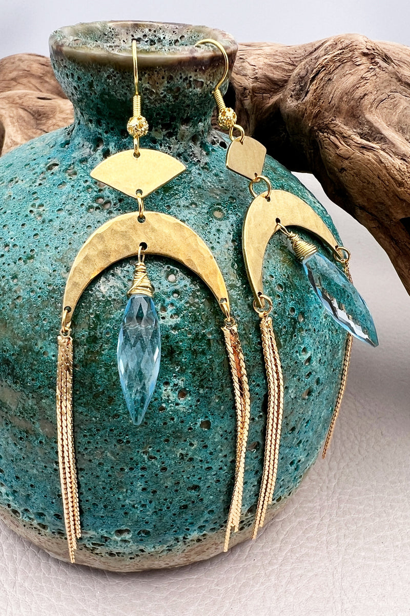 Gold statement earrings with a faceted aqua crystal, a celestial moon charm, and flowing golden tassels. Bold, lightweight, and designed for movement and shine.