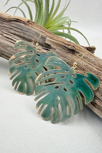 A pair of gold-tone dangle earrings shaped like green monstera leaves, displayed on a piece of driftwood with a small air plant in the background.