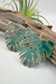 A pair of gold-tone dangle earrings shaped like green monstera leaves, displayed on a piece of driftwood with a small air plant in the background.