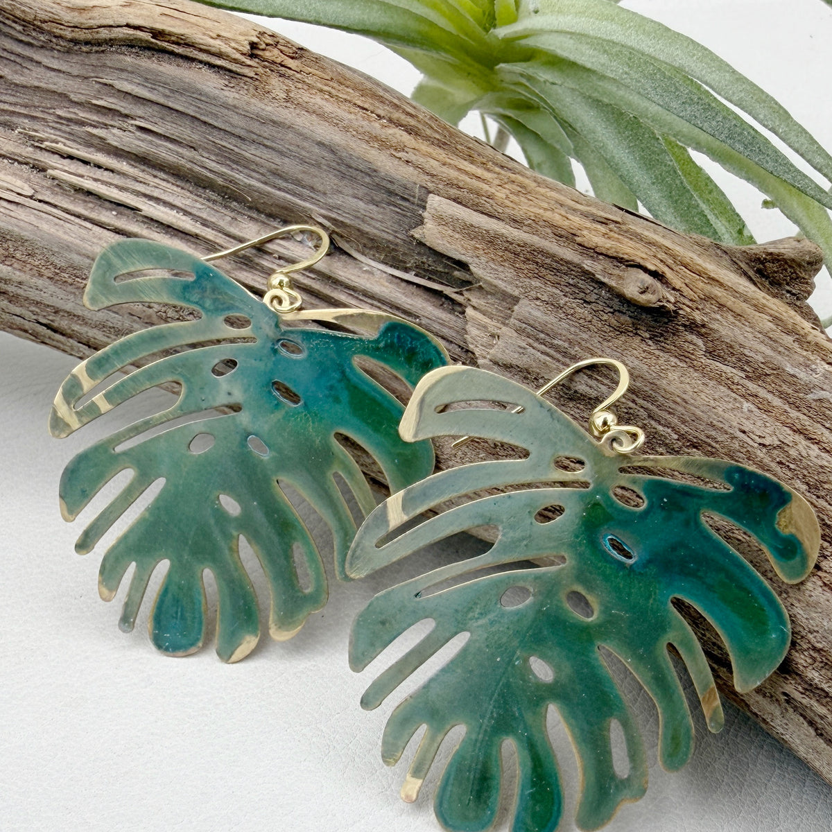 A pair of dangling earrings shaped like monstera leaves, with a translucent green and teal finish and gold-tone hooks, displayed on a piece of driftwood with a small green plant in the background.