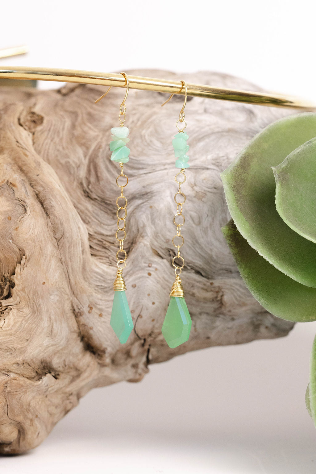 Green teardrop earrings with gold hooks on a wooden surface with a succulent plant.