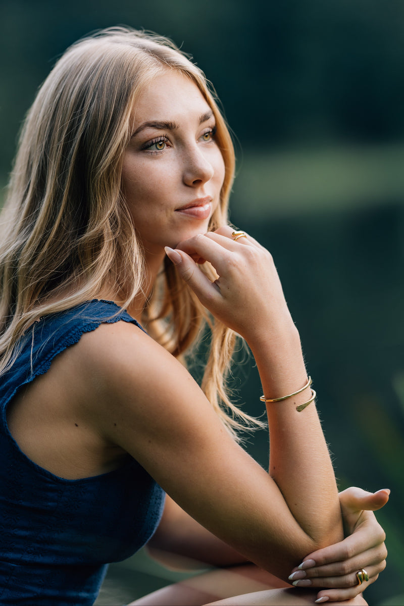 teenager girl in a blue dress sitting outdoors with a blurred green background wearing a hammered twisted golden bracelet