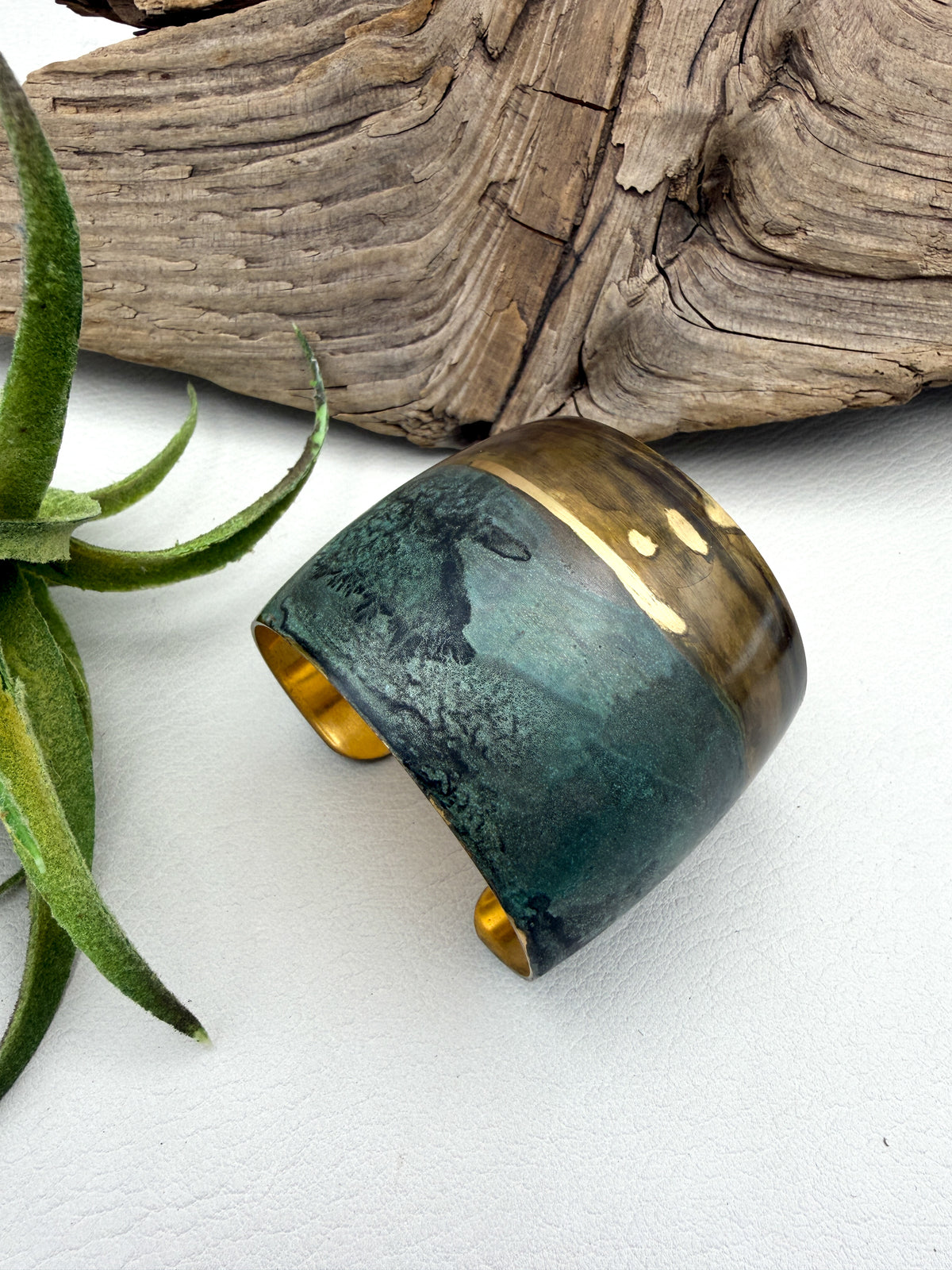 A wide patina bracelet with a distressed teal and dark green finish and polished gold-toned ends, styled on a white surface with driftwood and a green plant in the background.