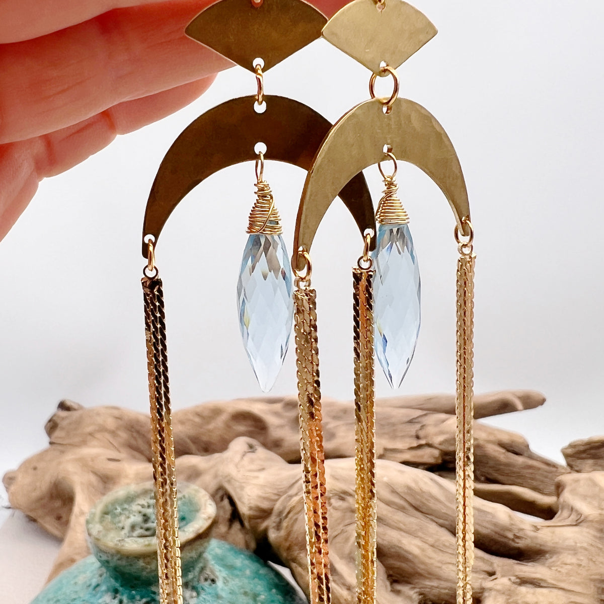Gold statement earrings with a faceted aqua crystal, a celestial moon charm, and flowing golden tassels. Bold, lightweight, and designed for movement and shine.