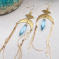 Gold statement earrings with a faceted aqua crystal, a celestial moon charm, and flowing golden tassels. Bold, lightweight, and designed for movement and shine.